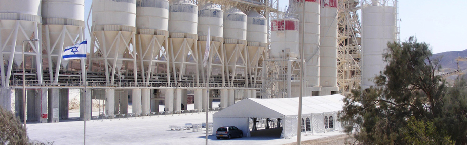 מתקן תעשייתי חיצוני עם מספר מכלי אחסון גליליים לבנים וגבוהים הנתמכים במסגרות מתכת. בחזית נמצא אוהל לבן גדול ומתחתיו רכב חונה. משמאל נראה דגל ישראל, וברקע גבעות מדבריות ועצים דלילים תחת שמיים בהירים.