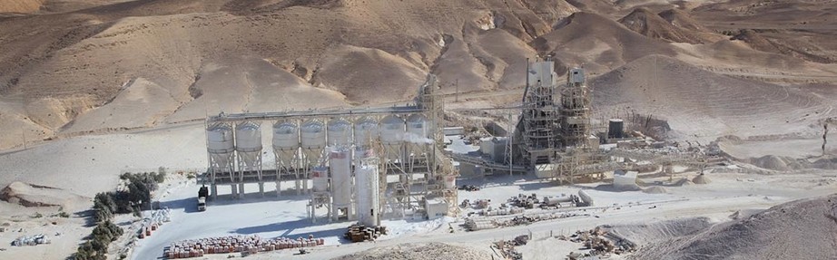 מבט אווירי רחב על מתקן תעשייתי גדול בנוף מדברי. האתר כולל מספר מכלי אחסון גליליים לבנים וגבוהים, מסגרות מתכת, מתקני עיבוד ומערכות מסועים הפרוסים על שטח מישורי. סביב המתקן גבעות סלעיות חשופות, חומרי בנייה וכלי רכב הנראים על הקרקע באור יום.