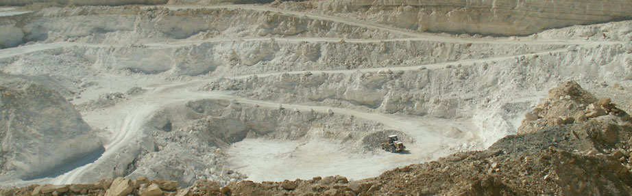 מבט רחב על מחצבה פתוחה עם קירות סלע מדורגים החצובים באבן בהירה. דרכי עפר מתפתלות יורדות לתוך המחצבה, ובמרכז פועל כלי חפירה קטן. בחזית נראים סלעים וחצץ, ללא צמחייה.