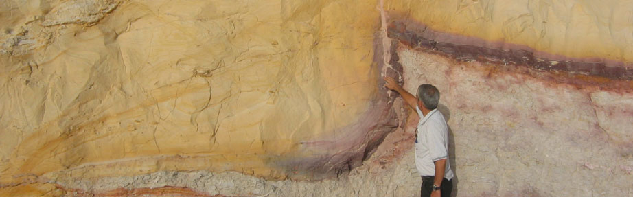 אדם עומד לצד קיר סלע חשוף במחצבה ומצביע על שכבת מינרל אופקית מובחנת. קיר הסלע מציג שכבות צבעוניות בגוונים של צהוב, לבן ואדמדם המדגישות את המבנה הגאולוגי.