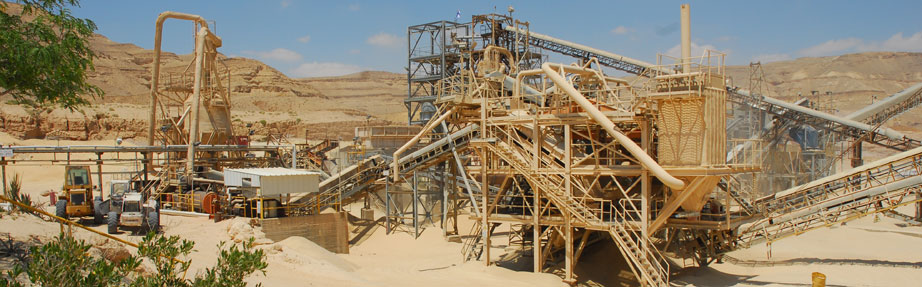 מתקן תעשייתי גדול לעיבוד מינרלים באזור מדברי, הכולל מבני מתכת מחוברים, מסועים, צינורות ומדרגות. ציוד מכני כבד פועל בגובה הקרקע, עם קרקע חולית וגבעות סלעיות ברקע תחת שמיים בהירים.