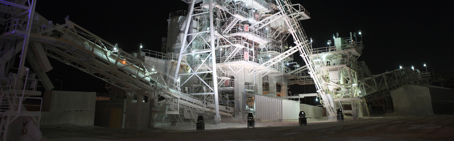 מבט לילי על מתקן עיבוד תעשייתי גדול המואר באורות חזקים. מסגרות מתכת גבוהות, מדרגות ומסועי חומרים יוצרים מבנה מורכב, עם מכלים וציוד הנראים על רקע שמיים כהים. תאורת קרקע מדגישה את המכונות וסביבת העבודה.