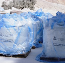 שקיות תעשייתיות גדולות, מלאות בחומר אבקה, מוערמות על משטחי עץ ועטופות בפלסטיק כחול שקוף. השקיות מאוחסנות בחוץ על משטח מכוסה אבקה לבנה, כשברקע נראית צמחייה דלילה.