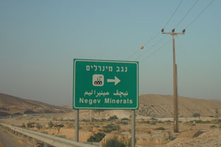 שלט דרכים באזור מדברי המציג את השם „נגב מינרלים” בעברית, בערבית ובאנגלית, עם גבעות ונוף פתוח ברקע.